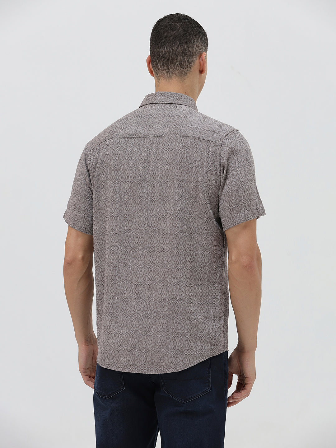 A man wearing a short-sleeve button-up shirt with a fine geometric pattern in gray and brown, paired with dark blue jeans, standing against a light gray studio background.