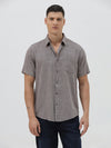 A man wearing a short-sleeve button-up shirt with a fine geometric pattern in gray and brown, paired with dark blue jeans, standing against a light gray studio background.