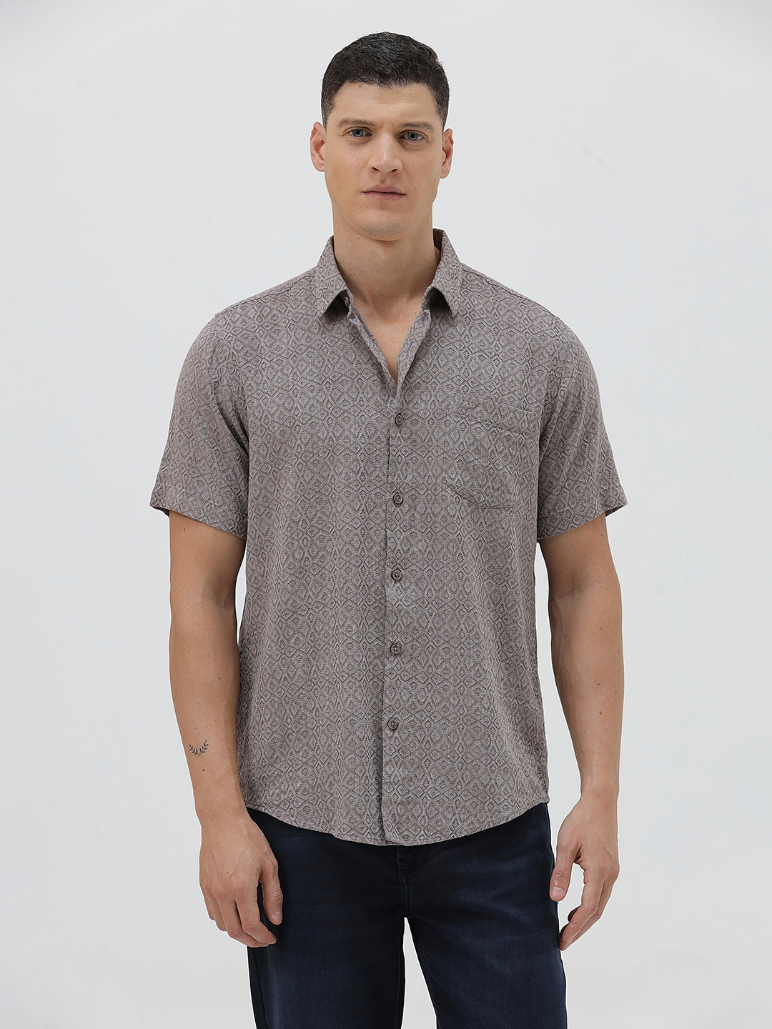 A man wearing a short-sleeve button-up shirt with a fine geometric pattern in gray and brown, paired with dark blue jeans, standing against a light gray studio background.