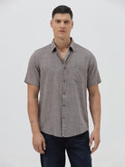 A man wearing a short-sleeve button-up shirt with a fine geometric pattern in gray and brown, paired with dark blue jeans, standing against a light gray studio background.