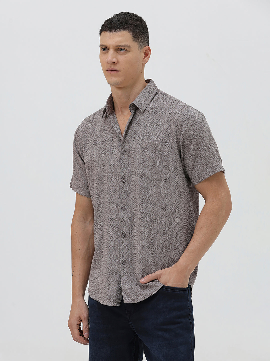 A man wearing a short-sleeve button-up shirt with a fine geometric pattern in gray and brown, paired with dark blue jeans, standing against a light gray studio background.