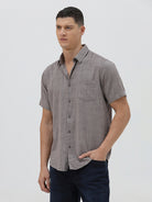 A man wearing a short-sleeve button-up shirt with a fine geometric pattern in gray and brown, paired with dark blue jeans, standing against a light gray studio background.