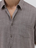 A man wearing a short-sleeve button-up shirt with a fine geometric pattern in gray and brown, paired with dark blue jeans, standing against a light gray studio background.