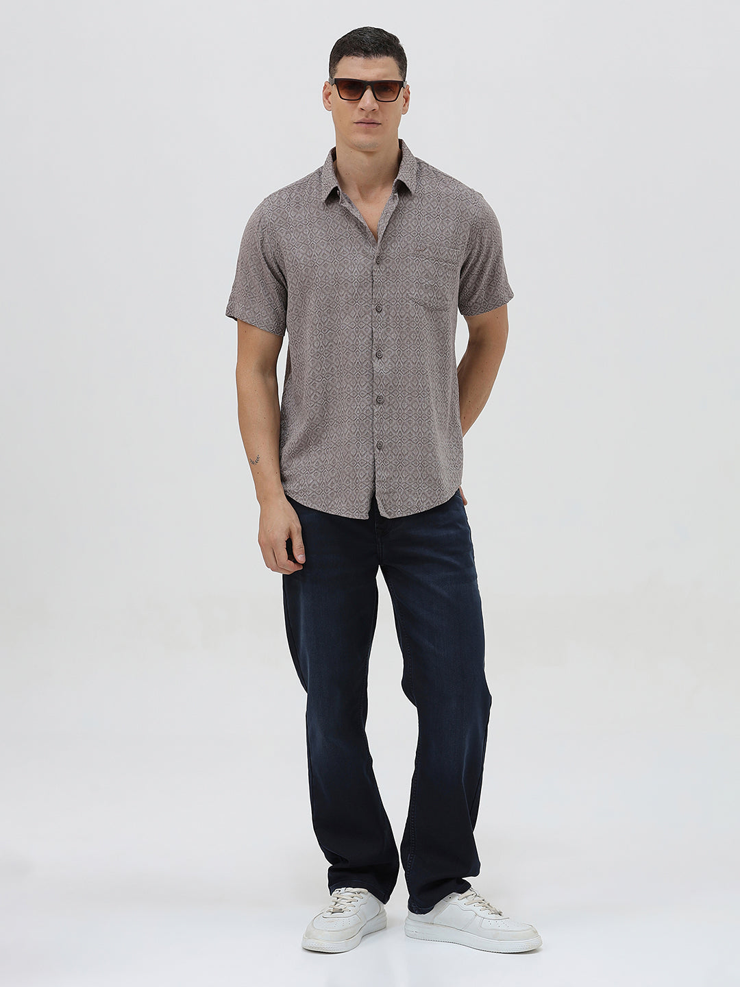 A man wearing a short-sleeve button-up shirt with a fine geometric pattern in gray and brown, paired with dark blue jeans, standing against a light gray studio background.
