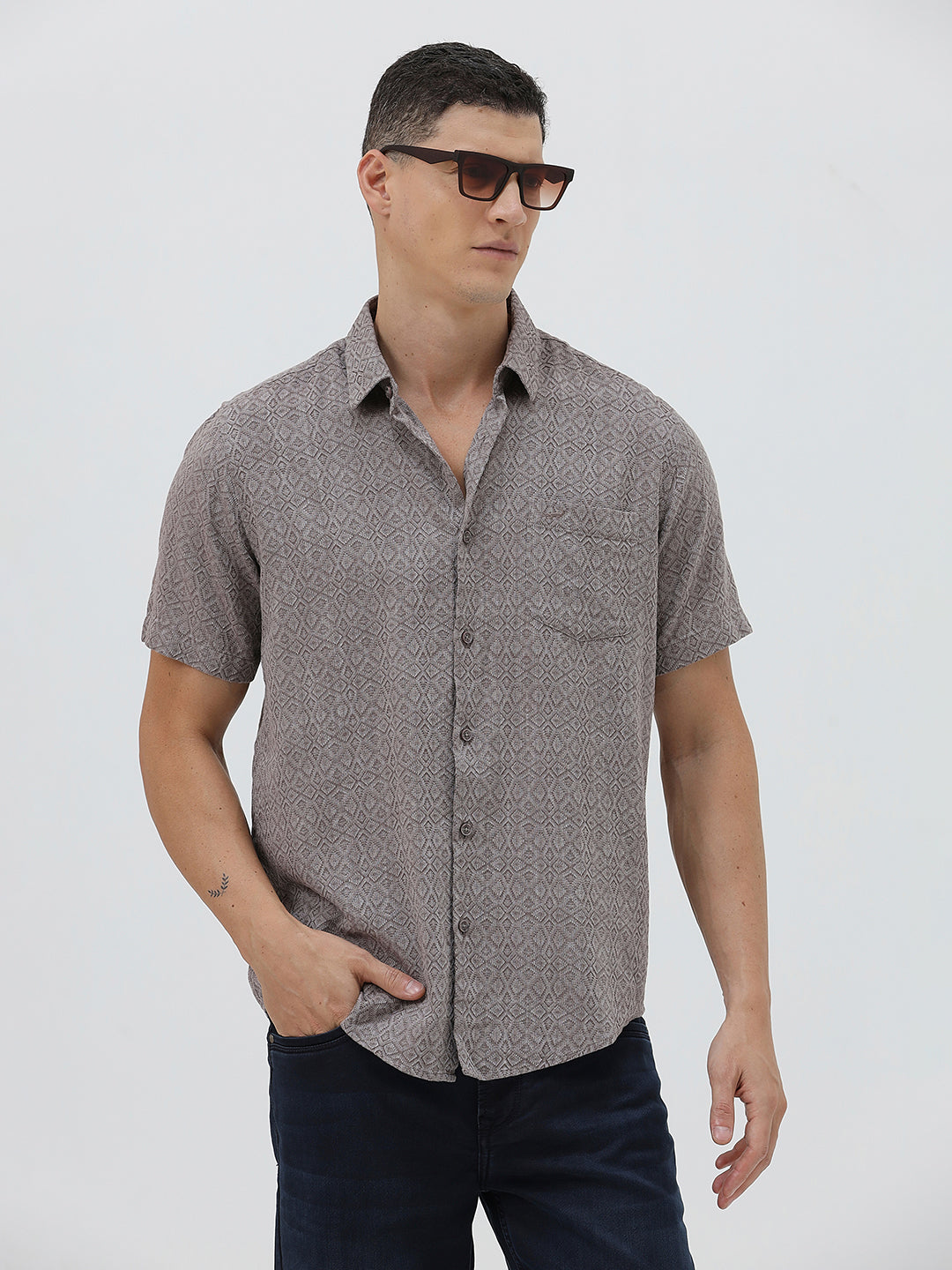 A man wearing a short-sleeve button-up shirt with a fine geometric pattern in gray and brown, paired with dark blue jeans, standing against a light gray studio background.