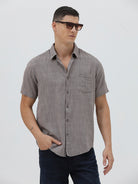 A man wearing a short-sleeve button-up shirt with a fine geometric pattern in gray and brown, paired with dark blue jeans, standing against a light gray studio background.