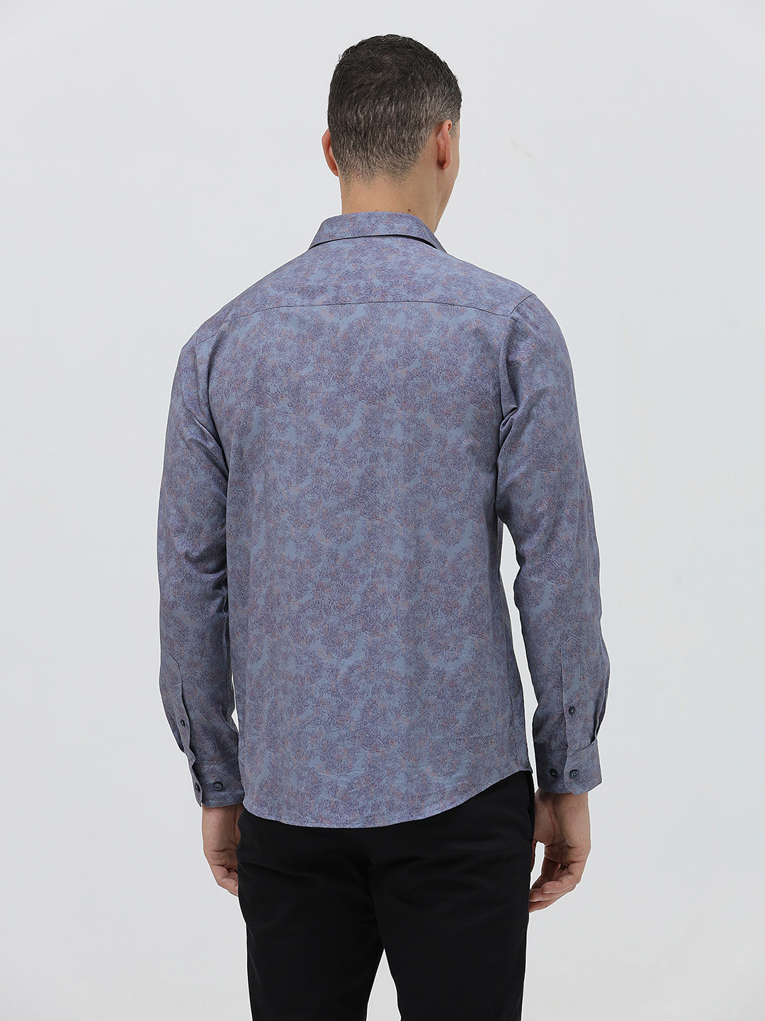 A man wearing a long-sleeve button-up shirt with a subtle blue and purple abstract pattern, paired with black trousers, standing against a light gray studio background.