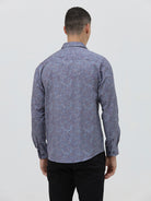 A man wearing a long-sleeve button-up shirt with a subtle blue and purple abstract pattern, paired with black trousers, standing against a light gray studio background.