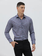 A man wearing a long-sleeve button-up shirt with a subtle blue and purple abstract pattern, paired with black trousers, standing against a light gray studio background.