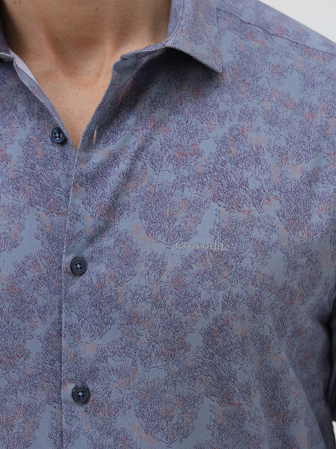 A man wearing a long-sleeve button-up shirt with a subtle blue and purple abstract pattern, paired with black trousers, standing against a light gray studio background.