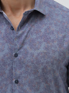 A man wearing a long-sleeve button-up shirt with a subtle blue and purple abstract pattern, paired with black trousers, standing against a light gray studio background.