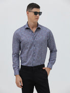 A man wearing a long-sleeve button-up shirt with a subtle blue and purple abstract pattern, paired with black trousers, standing against a light gray studio background.