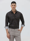 A man wearing a dark brown patterned long-sleeve button-down shirt with rolled-up sleeves and light gray trousers, standing against a light gray studio background.
