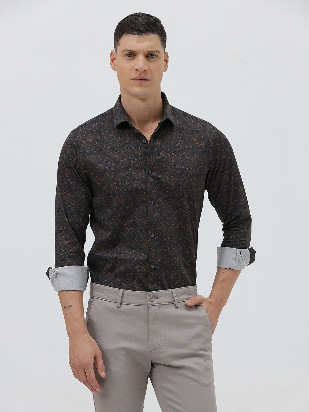 A man wearing a dark brown patterned long-sleeve button-down shirt with rolled-up sleeves and light gray trousers, standing against a light gray studio background.