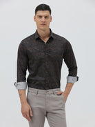 A man wearing a dark brown patterned long-sleeve button-down shirt with rolled-up sleeves and light gray trousers, standing against a light gray studio background.
