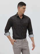 A man wearing a dark brown patterned long-sleeve button-down shirt with rolled-up sleeves and light gray trousers, standing against a light gray studio background.