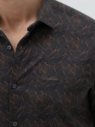 A man wearing a dark brown patterned long-sleeve button-down shirt with rolled-up sleeves and light gray trousers, standing against a light gray studio background.
