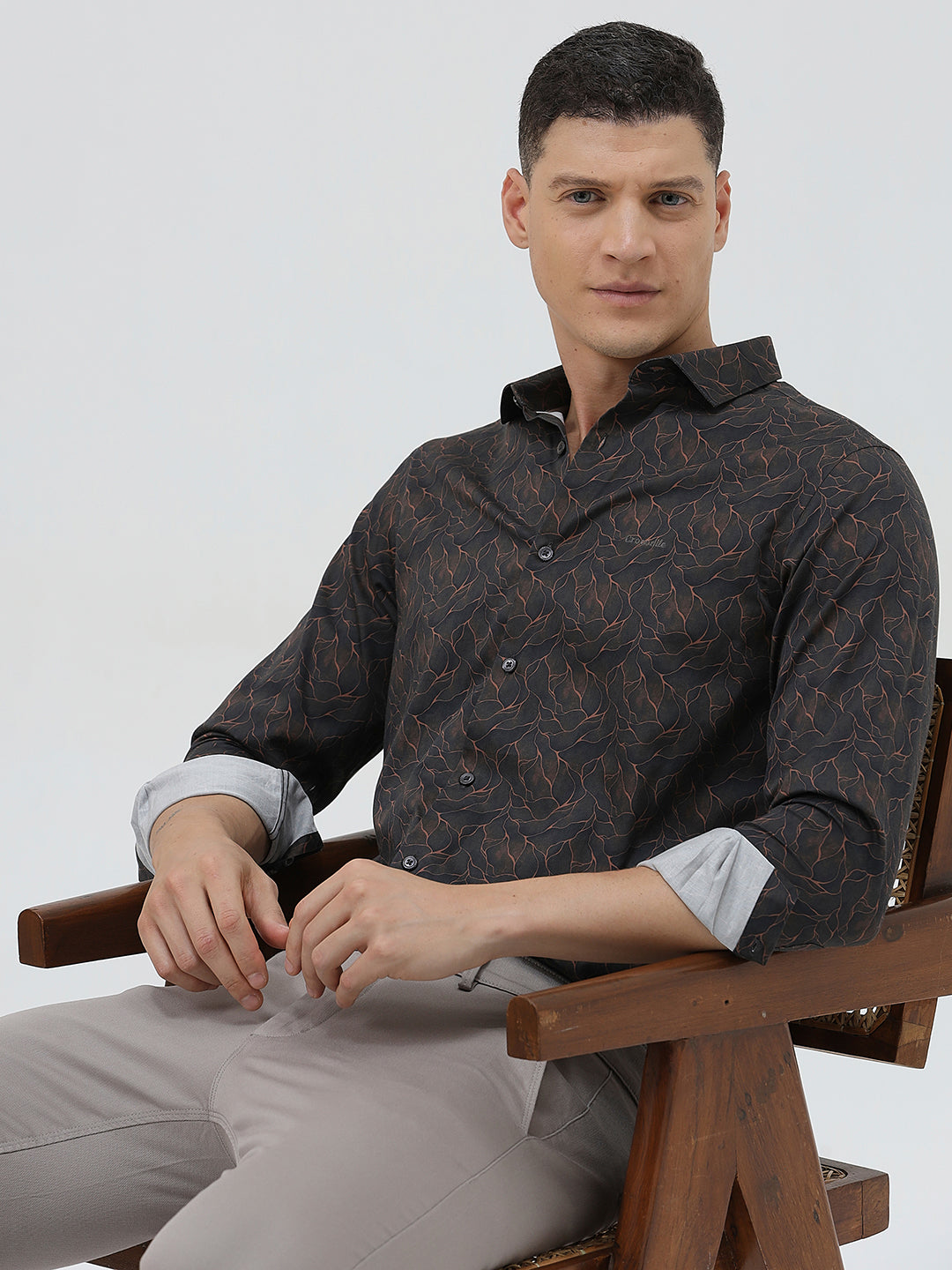 A man wearing a dark brown patterned long-sleeve button-down shirt with rolled-up sleeves and light gray trousers, standing against a light gray studio background.