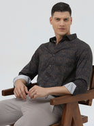 A man wearing a dark brown patterned long-sleeve button-down shirt with rolled-up sleeves and light gray trousers, standing against a light gray studio background.