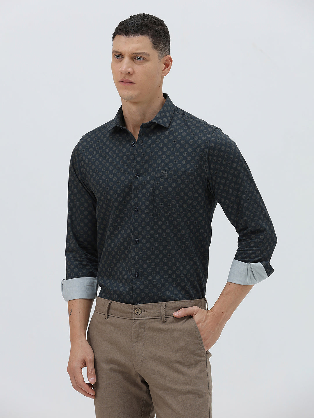 A man wearing a dark navy long-sleeve button-up shirt with a small geometric pattern and rolled-up sleeves, paired with khaki pants, against a plain light gray background.