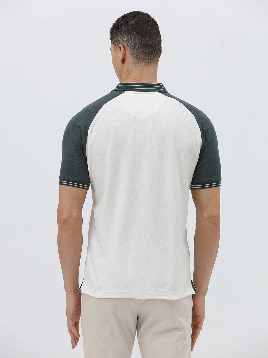 A man modeling a short-sleeve polo shirt with a white body and dark green raglan sleeves, featuring a striped collar and a small 'CR' logo on the chest, paired with beige trousers against a light gray background.