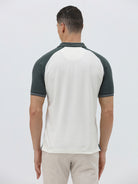 A man modeling a short-sleeve polo shirt with a white body and dark green raglan sleeves, featuring a striped collar and a small 'CR' logo on the chest, paired with beige trousers against a light gray background.