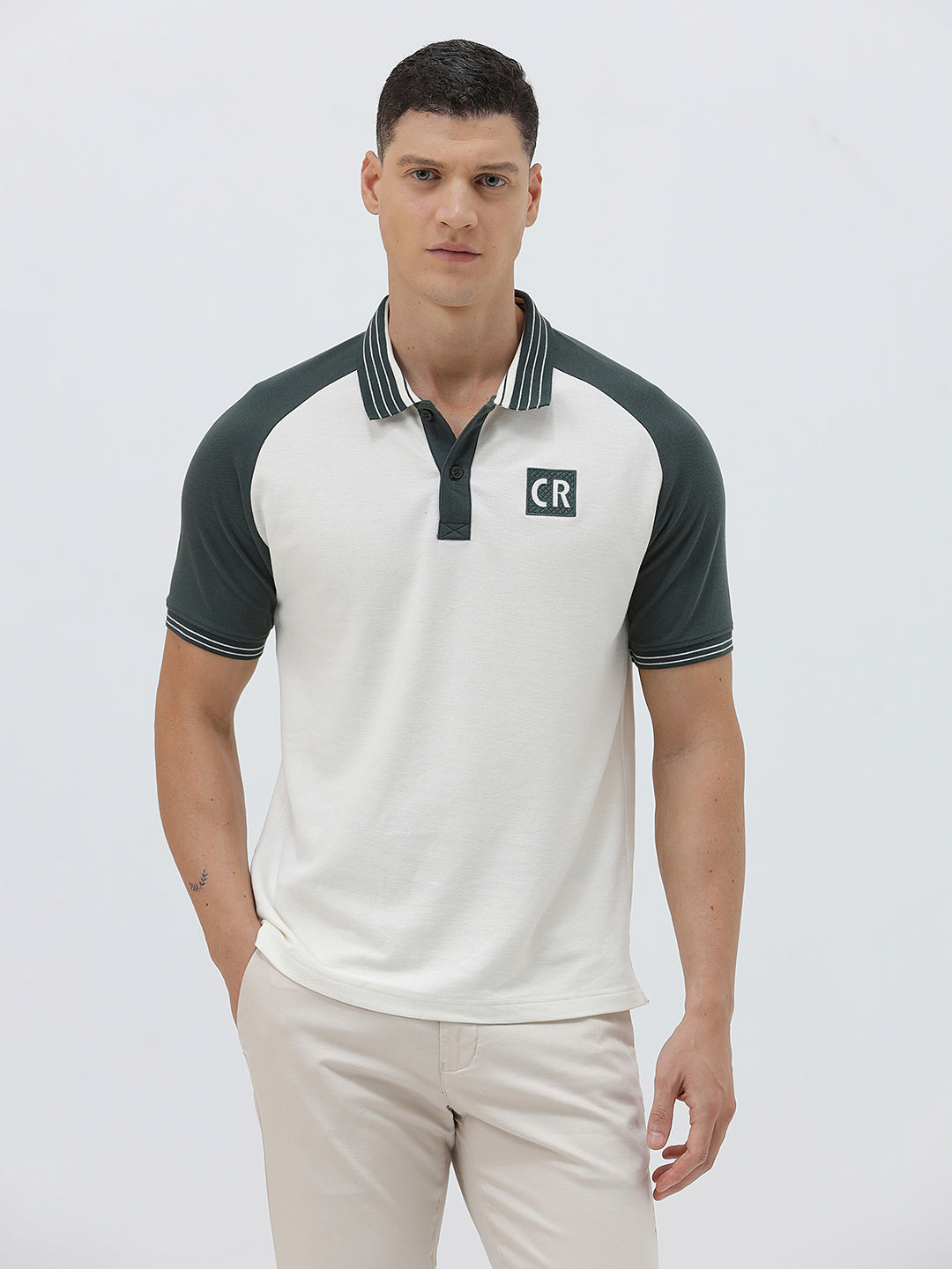 A man modeling a short-sleeve polo shirt with a white body and dark green raglan sleeves, featuring a striped collar and a small 'CR' logo on the chest, paired with beige trousers against a light gray background.