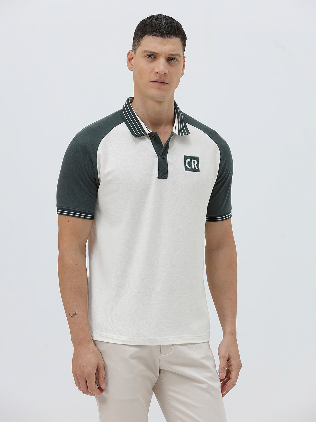 A man modeling a short-sleeve polo shirt with a white body and dark green raglan sleeves, featuring a striped collar and a small 'CR' logo on the chest, paired with beige trousers against a light gray background.