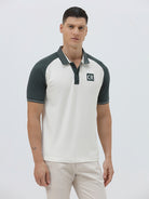 A man modeling a short-sleeve polo shirt with a white body and dark green raglan sleeves, featuring a striped collar and a small 'CR' logo on the chest, paired with beige trousers against a light gray background.