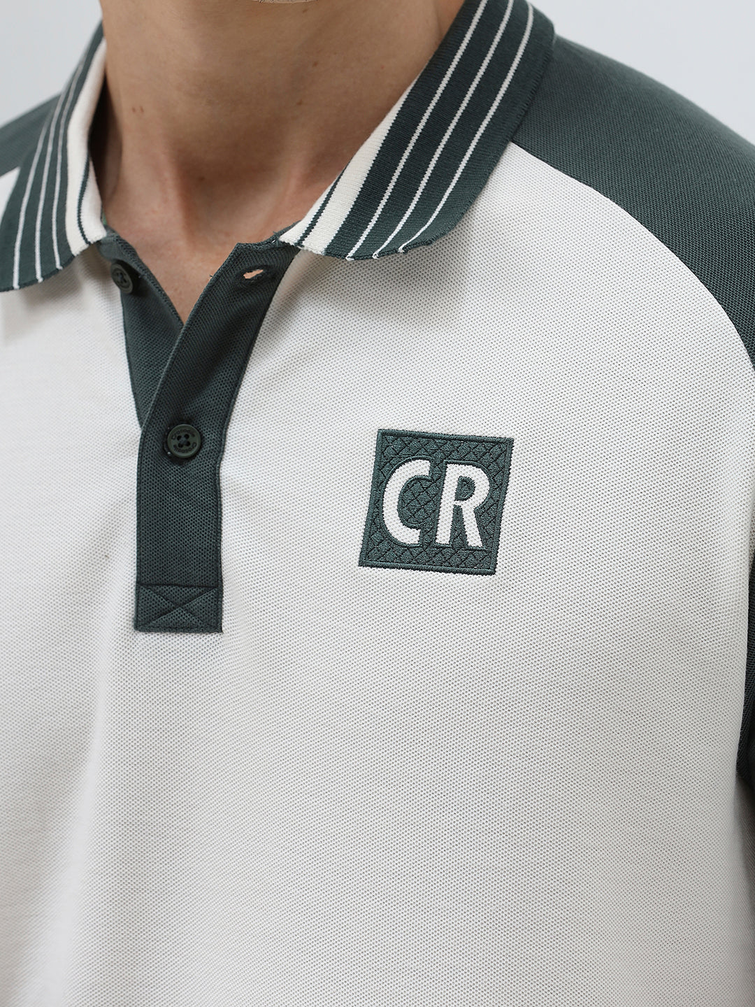 A man modeling a short-sleeve polo shirt with a white body and dark green raglan sleeves, featuring a striped collar and a small 'CR' logo on the chest, paired with beige trousers against a light gray background.