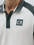 A man modeling a short-sleeve polo shirt with a white body and dark green raglan sleeves, featuring a striped collar and a small 'CR' logo on the chest, paired with beige trousers against a light gray background.