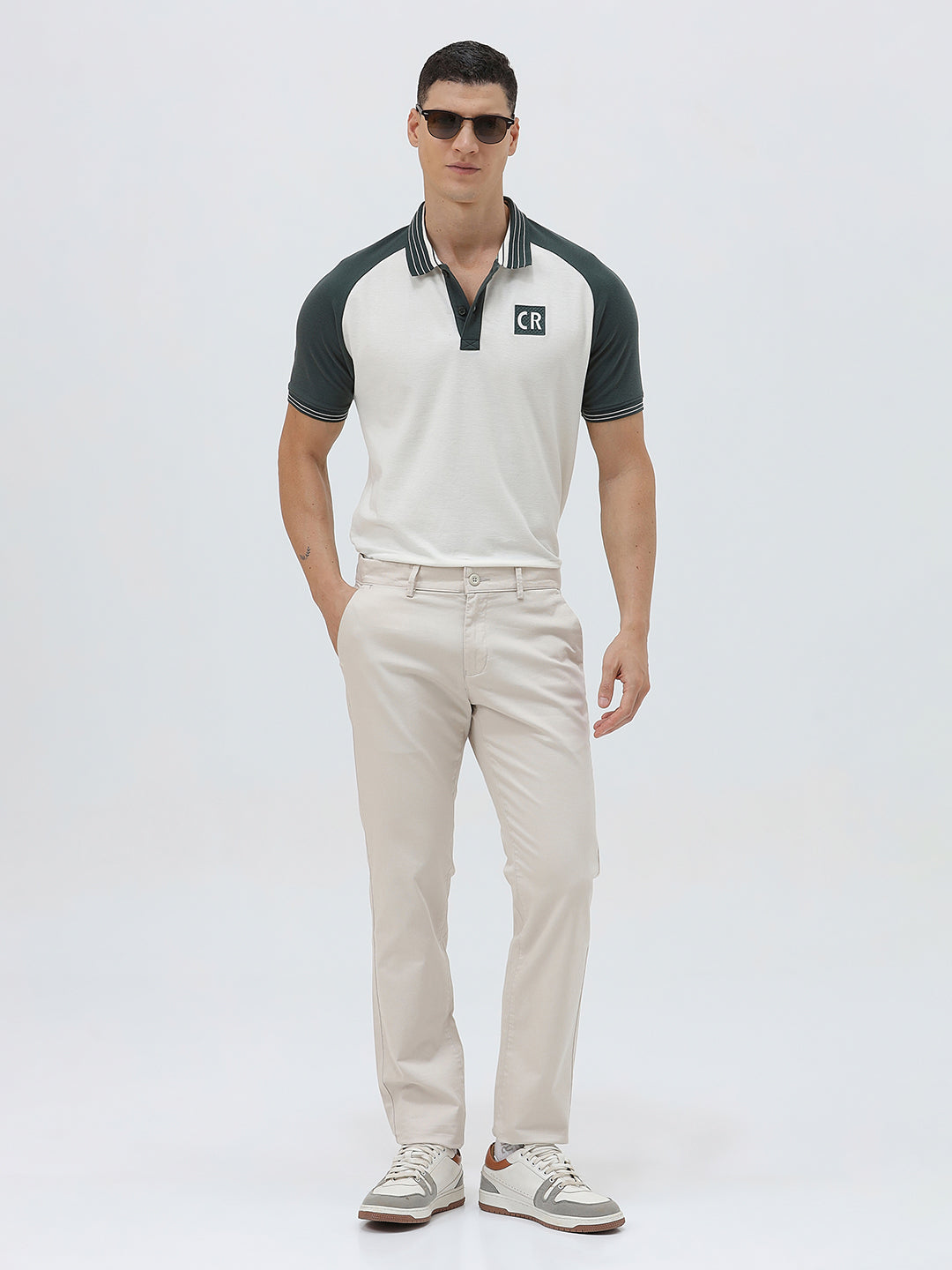 A man modeling a short-sleeve polo shirt with a white body and dark green raglan sleeves, featuring a striped collar and a small 'CR' logo on the chest, paired with beige trousers against a light gray background.