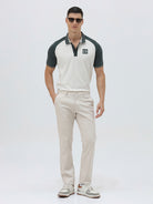 A man modeling a short-sleeve polo shirt with a white body and dark green raglan sleeves, featuring a striped collar and a small 'CR' logo on the chest, paired with beige trousers against a light gray background.