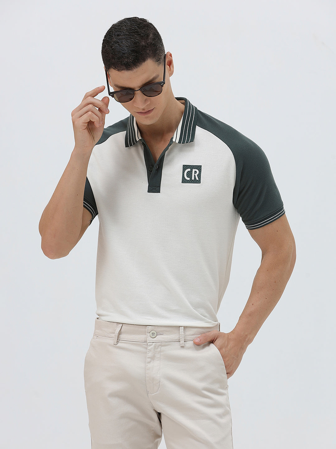 A man modeling a short-sleeve polo shirt with a white body and dark green raglan sleeves, featuring a striped collar and a small 'CR' logo on the chest, paired with beige trousers against a light gray background.