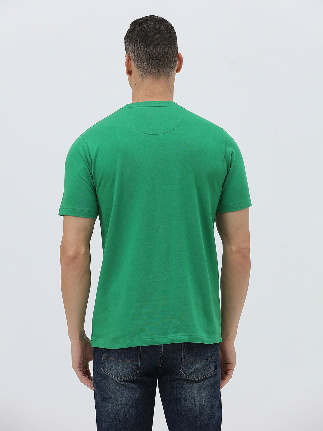 A man wearing a bright green crew-neck t-shirt with a small gold-toned graphic on the chest, paired with dark blue jeans, standing against a light gray studio background.