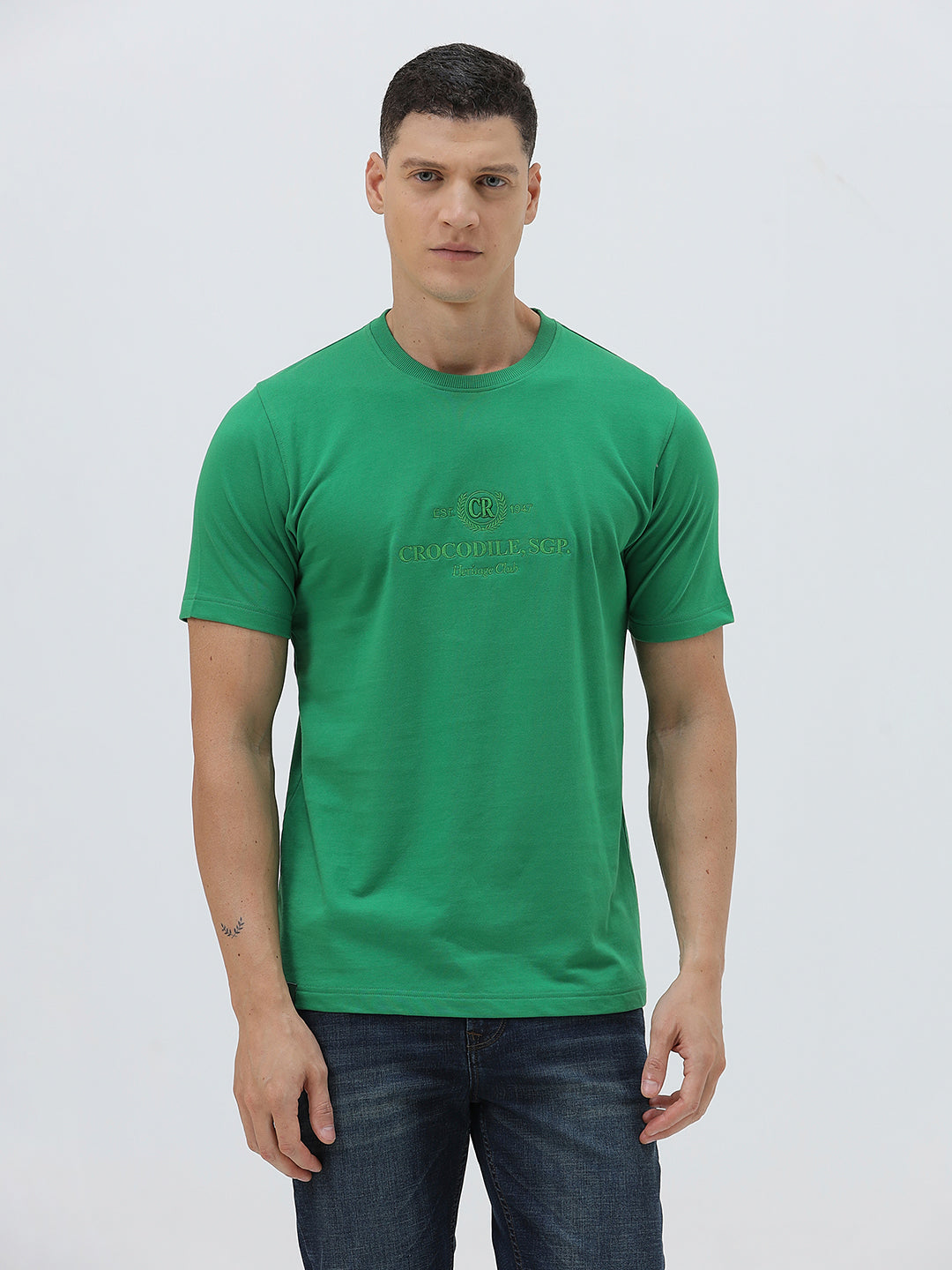 A man wearing a bright green crew-neck t-shirt with a small gold-toned graphic on the chest, paired with dark blue jeans, standing against a light gray studio background.