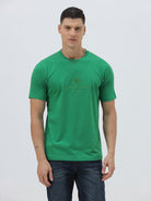 A man wearing a bright green crew-neck t-shirt with a small gold-toned graphic on the chest, paired with dark blue jeans, standing against a light gray studio background.