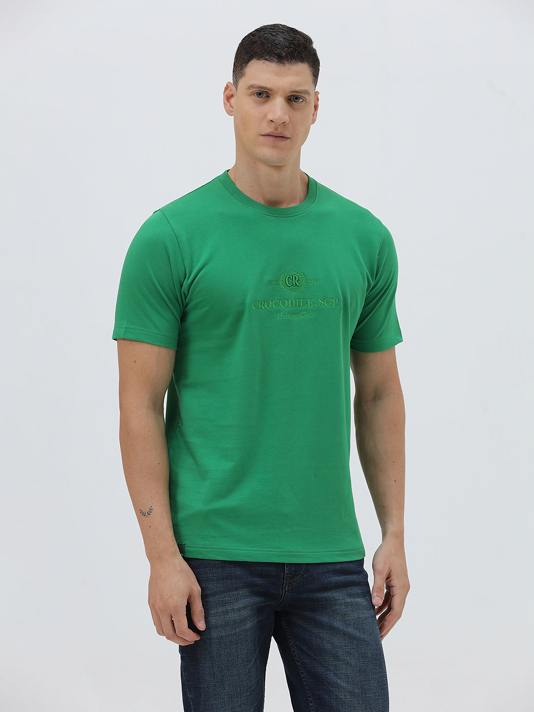 A man wearing a bright green crew-neck t-shirt with a small gold-toned graphic on the chest, paired with dark blue jeans, standing against a light gray studio background.