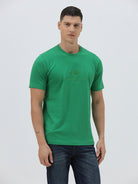 A man wearing a bright green crew-neck t-shirt with a small gold-toned graphic on the chest, paired with dark blue jeans, standing against a light gray studio background.