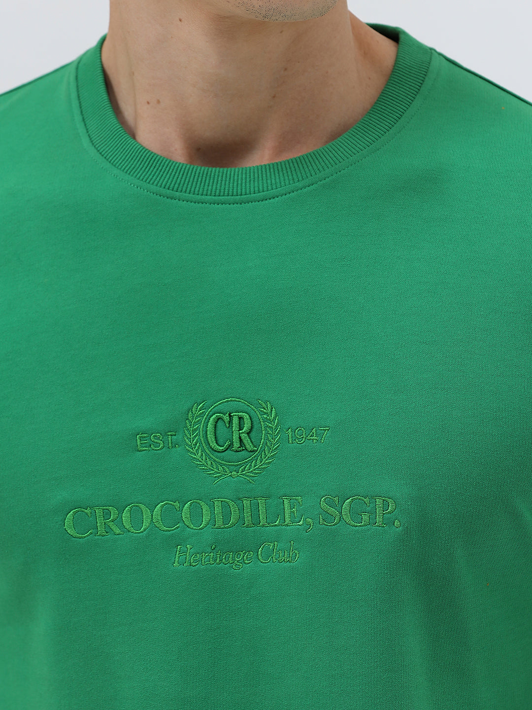 A man wearing a bright green crew-neck t-shirt with a small gold-toned graphic on the chest, paired with dark blue jeans, standing against a light gray studio background.