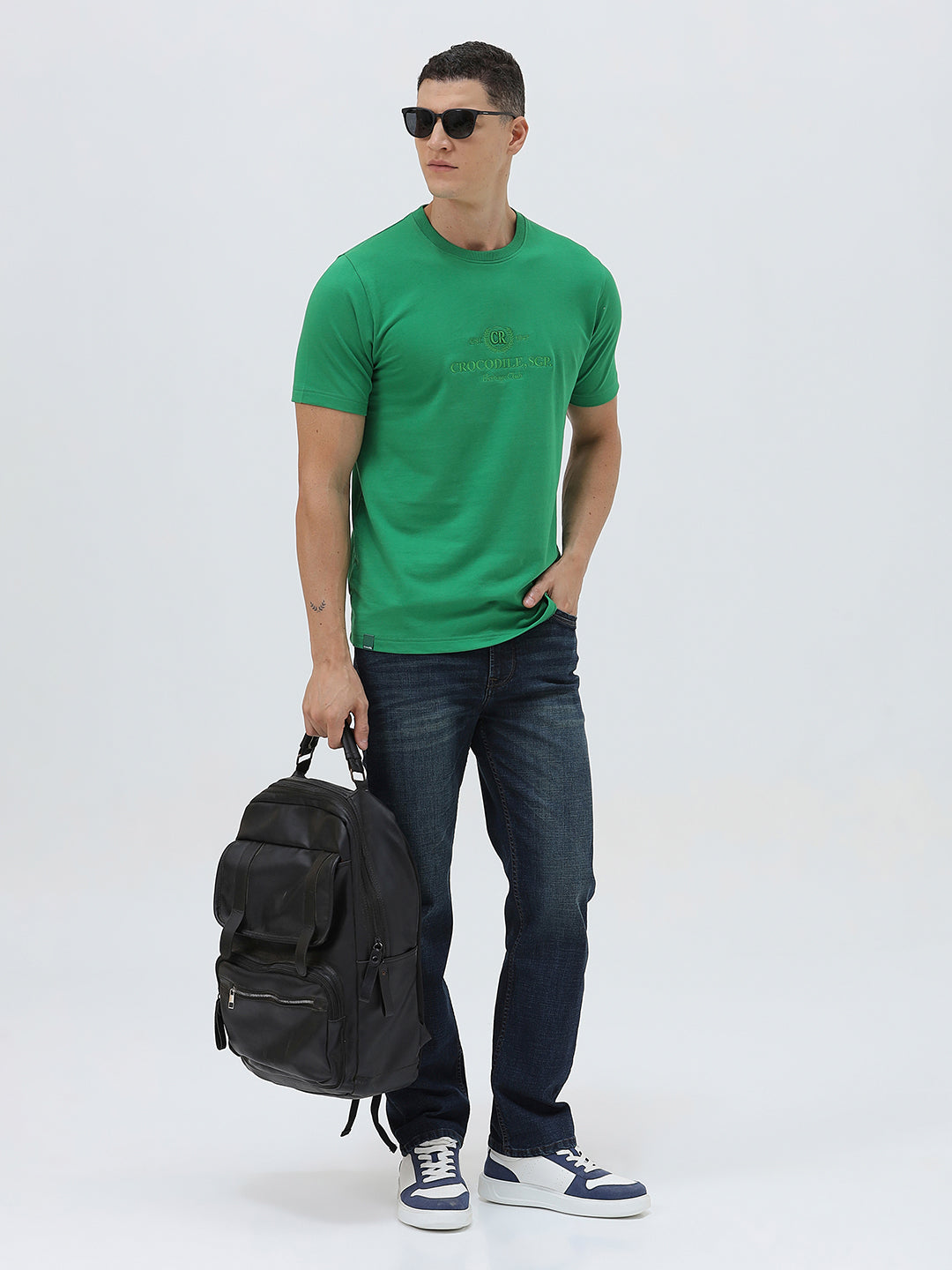 A man wearing a bright green crew-neck t-shirt with a small gold-toned graphic on the chest, paired with dark blue jeans, standing against a light gray studio background.