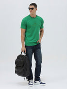 A man wearing a bright green crew-neck t-shirt with a small gold-toned graphic on the chest, paired with dark blue jeans, standing against a light gray studio background.