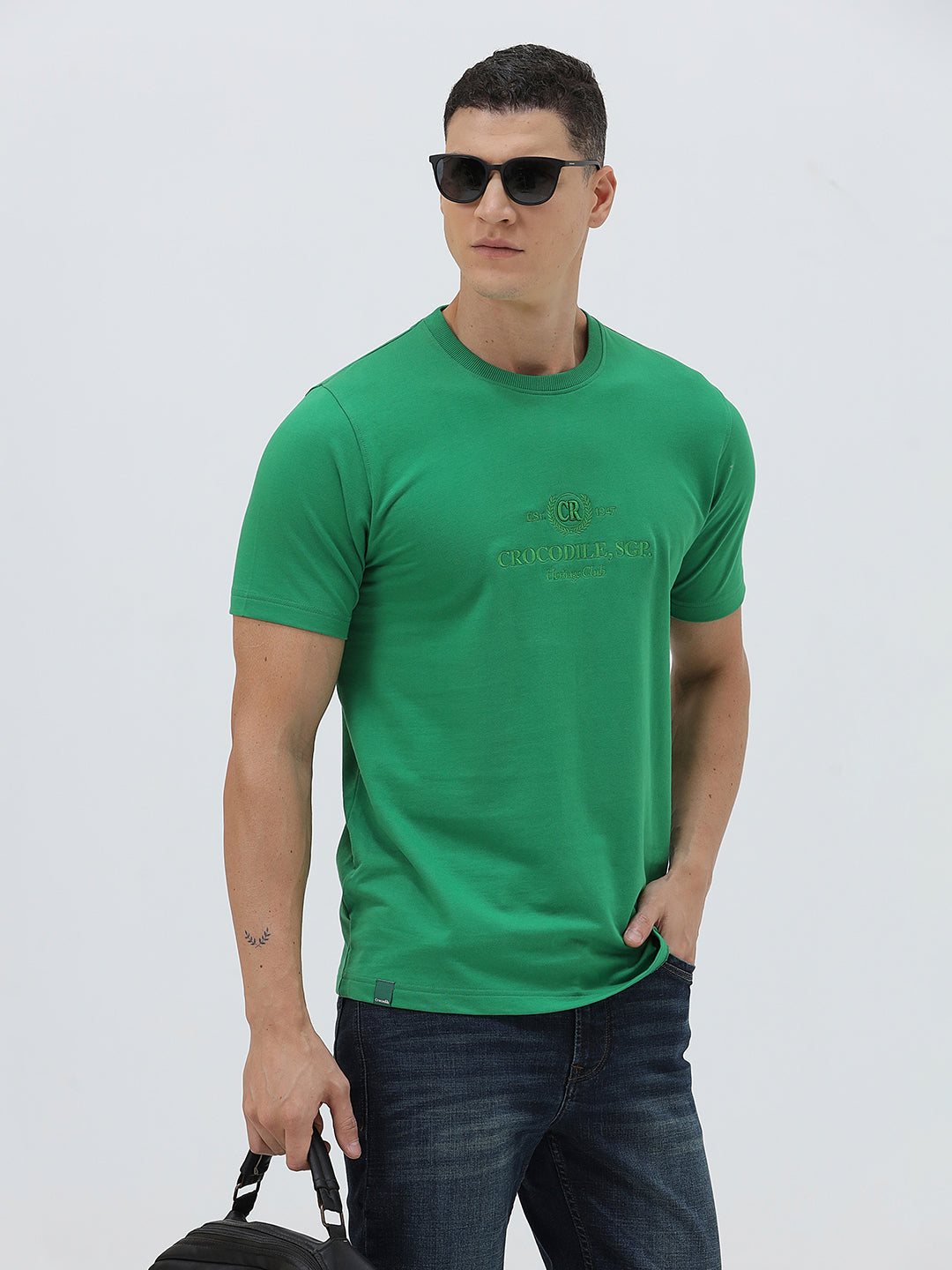 A man wearing a bright green crew-neck t-shirt with a small gold-toned graphic on the chest, paired with dark blue jeans, standing against a light gray studio background.