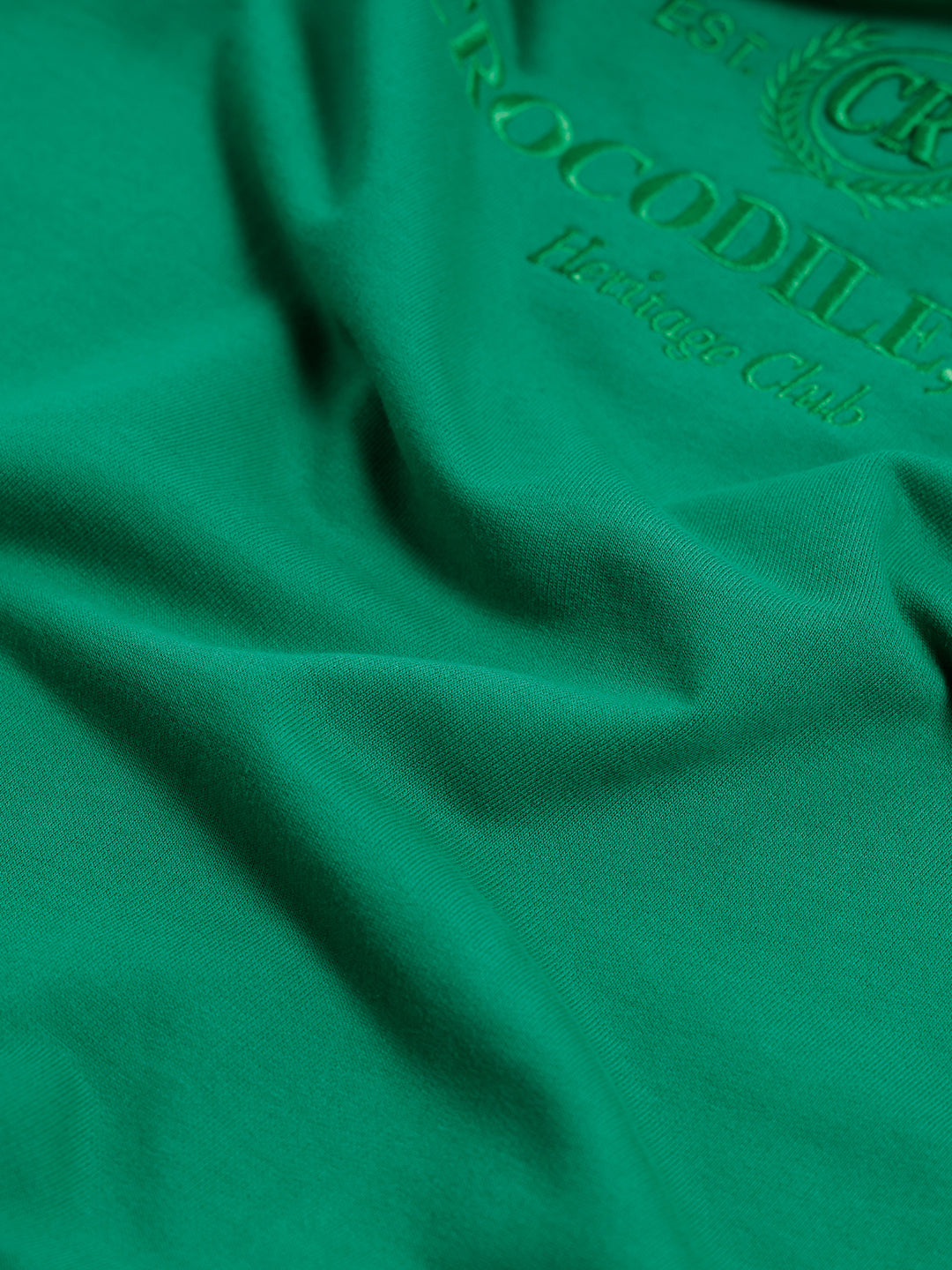 A man wearing a bright green crew-neck t-shirt with a small gold-toned graphic on the chest, paired with dark blue jeans, standing against a light gray studio background.