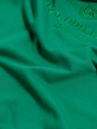 A man wearing a bright green crew-neck t-shirt with a small gold-toned graphic on the chest, paired with dark blue jeans, standing against a light gray studio background.