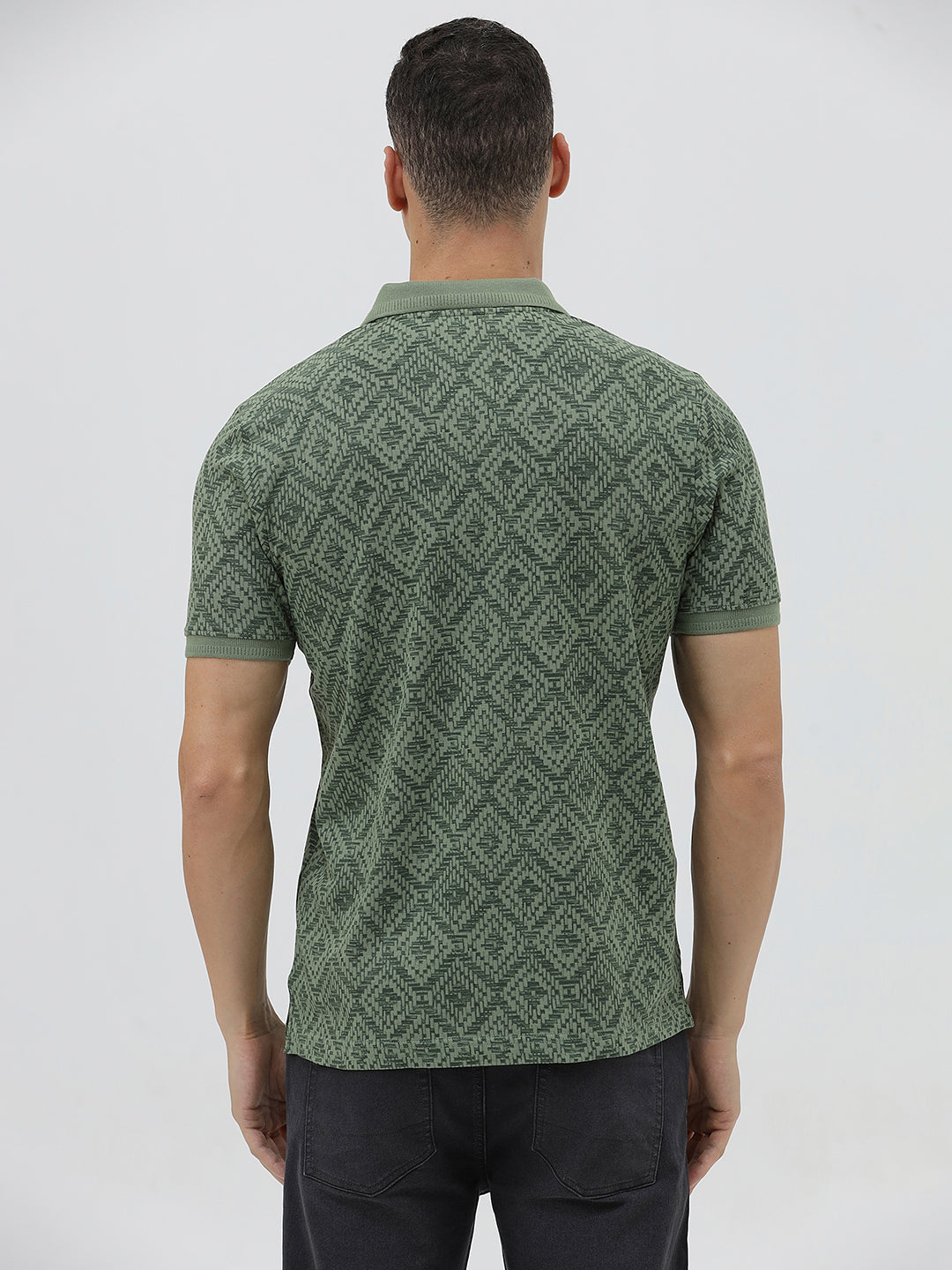 A man wearing a short-sleeve polo shirt with a green and dark gray geometric diamond pattern, paired with dark pants, standing against a light gray studio background.