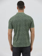 A man wearing a short-sleeve polo shirt with a green and dark gray geometric diamond pattern, paired with dark pants, standing against a light gray studio background.
