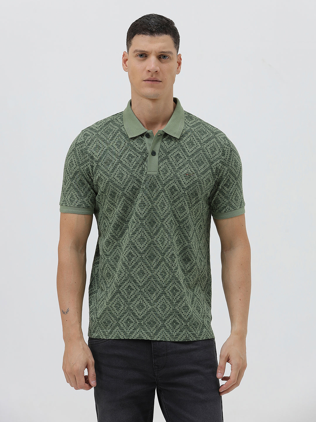 A man wearing a short-sleeve polo shirt with a green and dark gray geometric diamond pattern, paired with dark pants, standing against a light gray studio background.