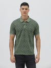 A man wearing a short-sleeve polo shirt with a green and dark gray geometric diamond pattern, paired with dark pants, standing against a light gray studio background.