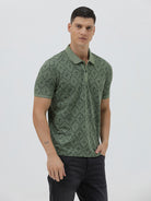 A man wearing a short-sleeve polo shirt with a green and dark gray geometric diamond pattern, paired with dark pants, standing against a light gray studio background.
